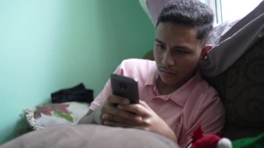 One hispanic man looking at phone screen sitting on couch at home. casual candid south american person using cellphone