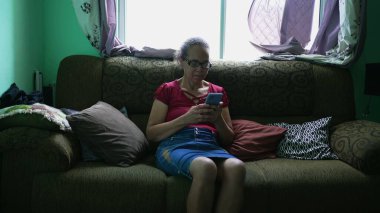 One candid hispanic senior woman using phone at home along sitting on couch