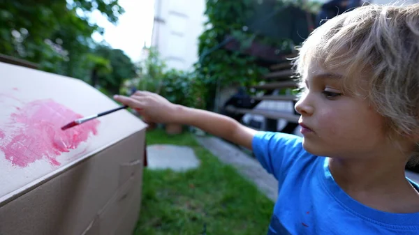 Artistic child painting outside in home garden. Artistic child playing with paint outside in garden painting cardboard box