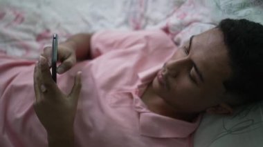 A young man lying in bed looking at smartphone device. A hispanic latin person using phone