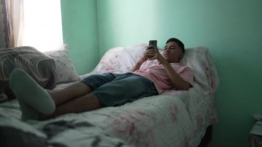 A young hispanic man looking at cellphone device in bedroom lying in bed