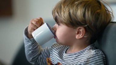 Toddler eating bread with jelly and drinking from mug in breakfast