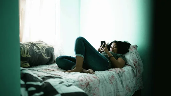 One black latina woman looking at cellphone lying in bedroom. A candid hispanic person using phone in bed