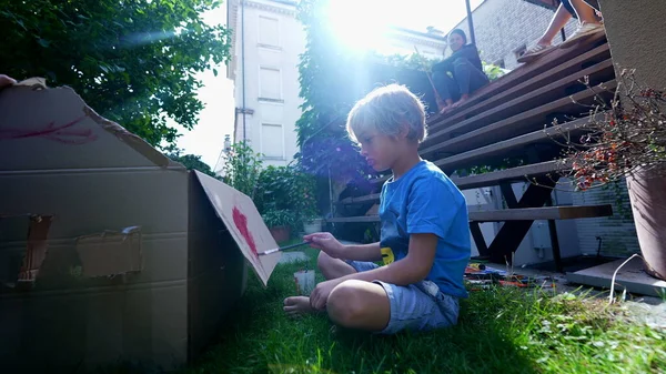 Artistic child painting outside in home garden. Artistic child playing with paint outside in garden painting cardboard box