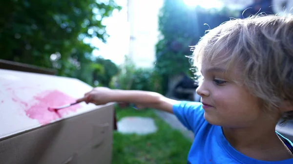 Artistic child painting outside in home garden. Artistic child playing with paint outside in garden painting cardboard box