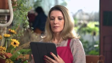 Elinde dijital tablet tutan, kırmızı önlük takan ve internetten siparişleri kontrol eden kadın işyeri sahibinin portresi.