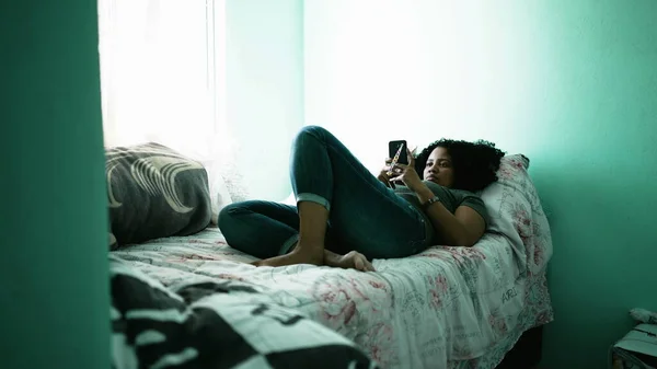 One black latina woman looking at cellphone lying in bedroom. A candid hispanic person using phone in bed