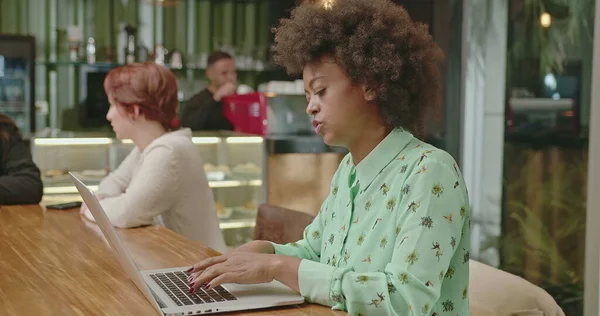 A happy black woman using laptop at coffee shop. One African American girl typing on computer keyboard smiling. A female entrepreneur person looking at laptop screen