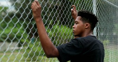 Pensive young black man leaning on metal fence. Contemplative mixed race person clipart