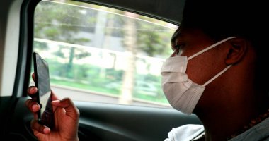 Black man in taxi browsing internet on smartphone device wearing pandemic mask