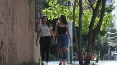 Lesbian girlfriends walking in city sidewalk outdoors. Two young women holding hands in slow-motion