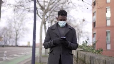 Siyah bir adam cep telefonuna bakarak dışarıda yürürken yüz maskesi takıyor.