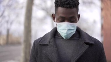 Siyah bir adam cep telefonuna bakarak dışarıda yürürken yüz maskesi takıyor.