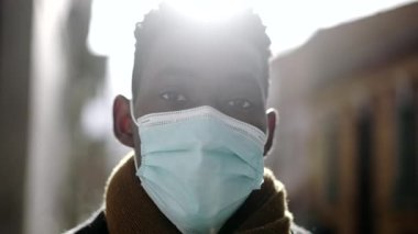 Siyahi bir adam güneş ışığında dışarıda kameraya bakarak maske takıyor.