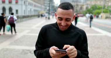Hispanic man using cellphone in street downtown, real people, latin guy browsing smartphone