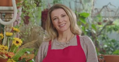 Businesswoman owner of small flower shop standing inside retail store smiling at camera while employees work in the background. Small business concept of female entrepreneur starting new business