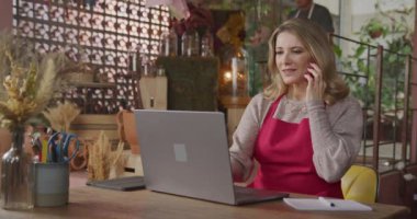 Owner of small business taking online orders in front of laptop computer. Female entrepreneur of local flower shop talking with customer or partner online wearing apron talking on phone