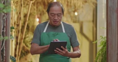 Small business owner checking inventory holding tablet and wearing apron. Asian American entrepreneur standing inside retail store shop