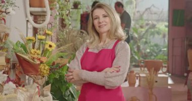 Businesswoman owner of small flower shop standing inside retail store smiling at camera while employees work in the background. Small business concept of female entrepreneur starting new business