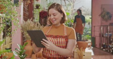 Female staff wearing apron using tablet device managing online orders inventory inside small business flower shop