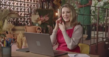 Small business owner working in front of computer managing online orders doing inventory talking on phone multi tasking. Flower delivery service. Female florist working online
