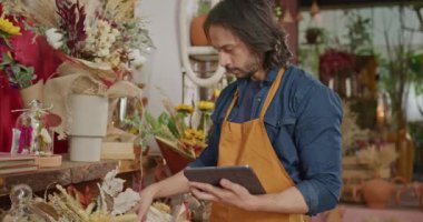 Employee of small business checking inventory holding tablet standing inside flower shop.  Male staff using modern technology to manage online orders