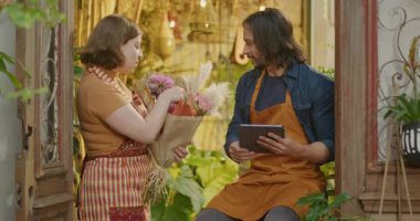 Two happy employees holding tablet and bouquet of flowers inside small local flower shop business store. Young male and female workers receiving online orders. Staff of retail store