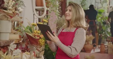 One happy middle aged female entrepreneur holding tablet standing inside flower shop checking for online orders with digital modern device. Small business owner checking inventory managing small business
