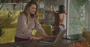 Woman entrepreneur owner of flower shop managing online orders with staff. Employee bringing bouquet of flowers for female boss pointing at online delivery for shipment inside floriculture store