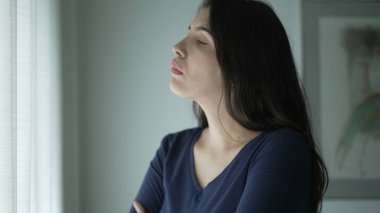Person standing by window with arms crossed consoling herself. Pensive thoughtful young woman