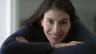 Portrait of woman in her 30s smiling at camera leaning on table with arms crossed closeup face