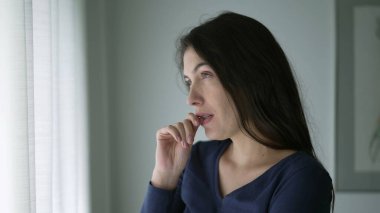 Anxious young woman trying to calm herself standing by window looking out. Person biting nail touching chest with hand