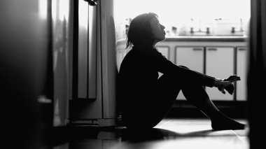 Depressed girl sitting on kitchen floor on monochrome. Black and white photography of woman in desperate moment