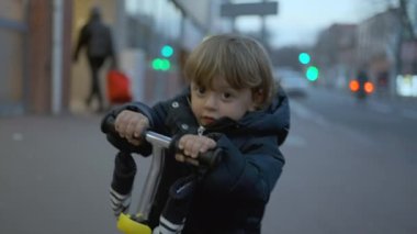 Little toddler boy riding three wheeled scooter in city at night during winter