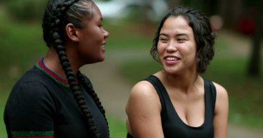 Two diverse friends laughing and smiling together. Mixed race girlfriends talking in conversation outside. Authentic real life laugh and smile