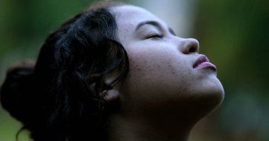 Yağmurda duran genç kadın gözleri kapalı gülümsüyor. Düşen damlaları hissederek hayattaki küçük şeylerden zevk alıyor.