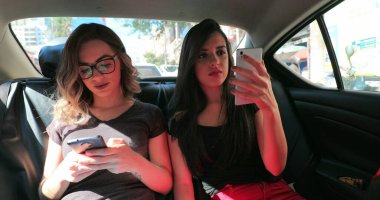 Candid friends in the back seat of a car checking their cellphone. Two girls looking at the smartphone