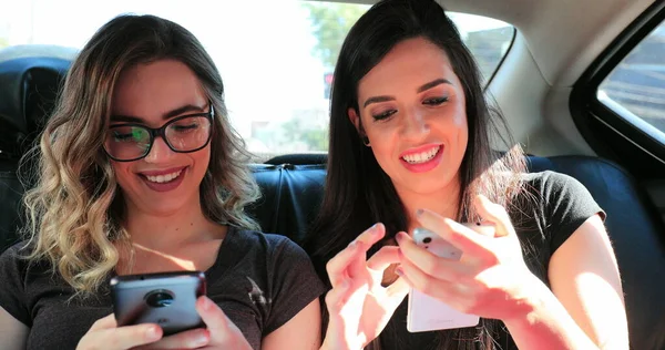 Candid friends holding cellphones in the back seat of a taxi Authentic girlfriends looking at smartphone real life moment