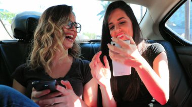 Friends real life laugh in the back seat of a car holding cellphone showing to friend content Candid girlfriends together laughing while riding taxi