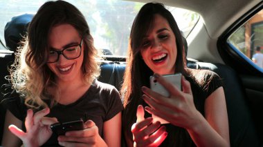 Female friends browsing on their smartphone while riding cab laughing. Women laughing while using cellphone in the back seat of a car