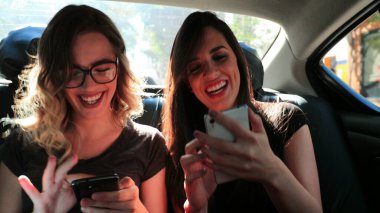 Female friends browsing on their smartphone while riding cab laughing. Women laughing while using cellphone in the back seat of a car