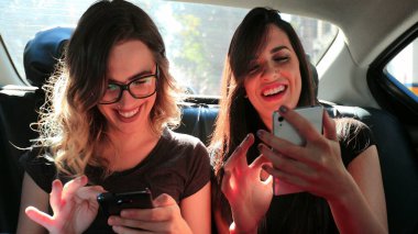 Female friends browsing on their smartphone while riding cab laughing. Women laughing while using cellphone in the back seat of a car