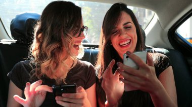 Female friends browsing on their smartphone while riding cab laughing. Women laughing while using cellphone in the back seat of a car