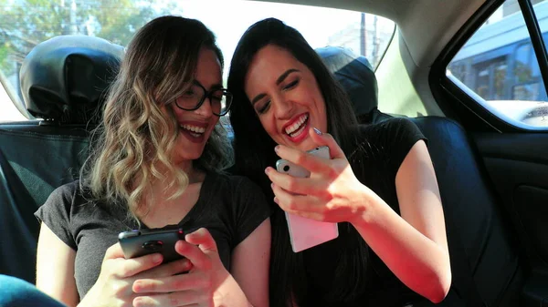 Friends real life laugh in the back seat of a car holding cellphone showing to friend content Candid girlfriends together laughing while riding taxi