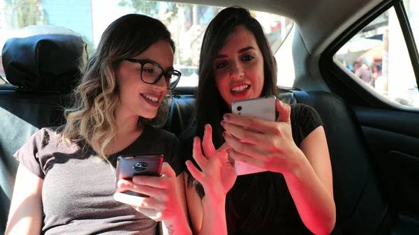 Friends looking at their cellphones in the back seat of a taxi cab gossiping and browsing the internet on their smartphones