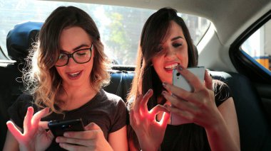 Female friends browsing on their smartphone while riding cab laughing. Women laughing while using cellphone in the back seat of a car