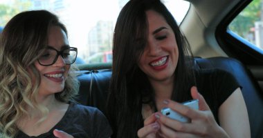 Candid girlfriends in the back seat of a car sharing cellphone screen showing content Friends together riding taxi cab holding smartphone