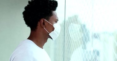 African american man wearing virus prevention mask looking out apartment window