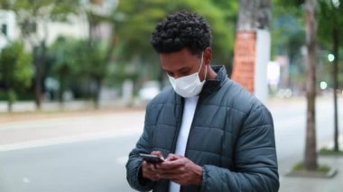 African american man looking at cellphone screen while walking in city downtown wearing covid-19 mask prevention