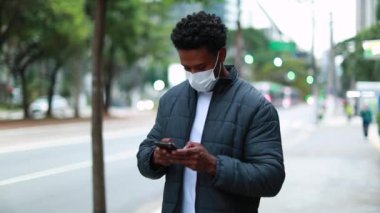 Black person checking smartphone while walking in city downtown wearing pandemic mask